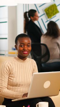 African american lady writing on laptop looking at camera smiling while diverse colleagues working in background. Multiethnic coworkers talking about startup financial company in modern business office