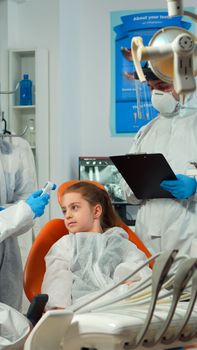 Stomatologist with face shield measuring girl temperature before dental examination during global pandemic. Concept of new normal dentist visit in coronavirus outbreak wearing protective suit.