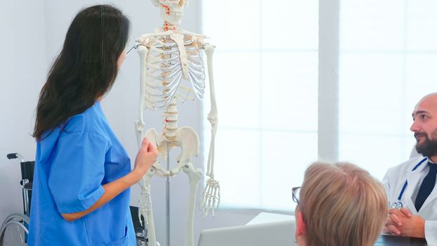 Female nurse demonstrating on skeleton in front of medical surgeons in conference room. Clinic expert therapist talking with colleagues about disease, medicine professional