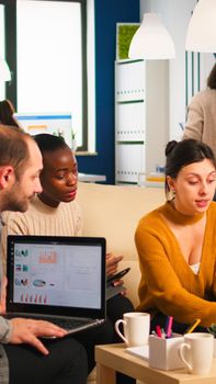 Businesswoman corporate leader talking to diverse team explaining project plan reviewing paperwork sitting on couch. Multiethnic coworkers taking notes on laptop and tablet analysing financial reports