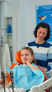 Child using finger to point affected tooth while the dentist talking with mother about oral toothache. Dentistry doctor explaining to mom the dental process, daughter sitting on stomatological chair
