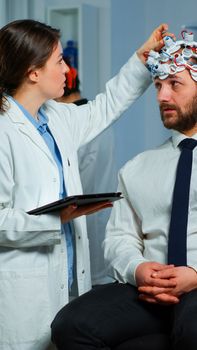 Man sitting on neurological chair with brainwave scanning headset, researcher doctor tracking evolution of patient's brain using tablet, adjusting eeg device. Scientist examining nervous system.