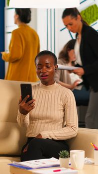 Tired black woman talking with colleague using video call holding smartphone with wiraless headset sitting on couch in business office. Diverse team planning new financial project in modern company