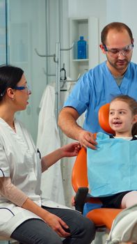 Little girl patient with tooth pain explaining dental problem to pediatric dentist and indicating at toothache with tongue. Stomatologist taking with mother about stomatologic examination of kid.