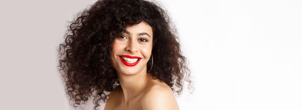 Close-up of cheerful smiling woman with red lipstick and curly hairstyle, looking happy, standing over white background.