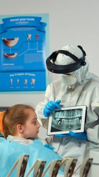 Dentist in protective equipment showing on tablet dental x-ray reviewing it with mother of kid patient. Medical team wearing face shield coverall, mask, gloves, explaining radiography using notebook