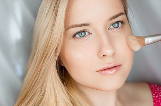 Beautiful blonde woman applying liquid make-up foundation on her skin with make-up brush.