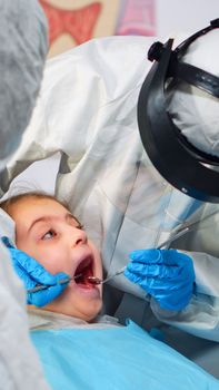 Close up of dentist in coverall examining girl patient in new normal stomatological office, standing near stomatological chair. Concept of doctor visit in coronavirus outbreak wearing face shield