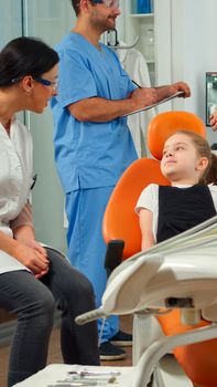 Nurse inviting kid patient in consultation dental room, pediatric doctor speaking with little girl. Man assistant talking with mother preparing for stomatologic examination in orthodontist office