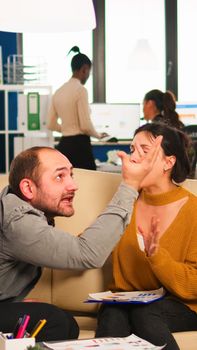 Angry man manager arguing woman employee for bad work result, sitting on couch, while diverse colleagues working scared in background. Business woman employee raging about multitasking difficult job.