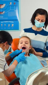 Stomatologist doctor checking tooth health of young patient using dental tools working in modern stomatological office with man nurse. Mother and daughter visiting doctor for periodic consulting