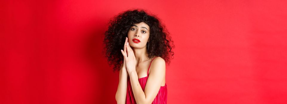 Romantic sensual woman with curly hair, wearing evening dress, posing seductive on red background.