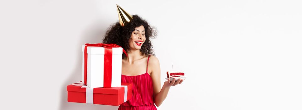 Cheerful smiling woman in red dress and party cone, looking happy at birthday cake, holding gifts, standing on white background. Copy space