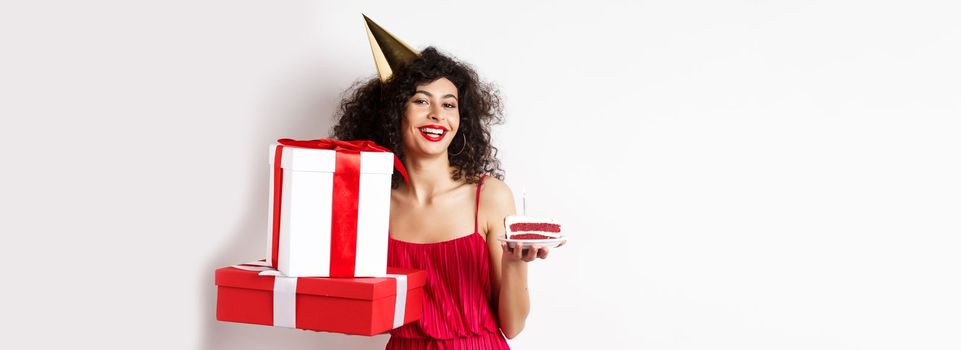 Happy birthday girl in red dress, celebrating and holding gifts with bday cake, standing on white background.