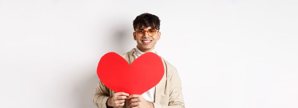 Handsome hipster guy with sunglasses and earring, waiting for true love on Valentines day, holding big red heart and smiling, standing over white background.