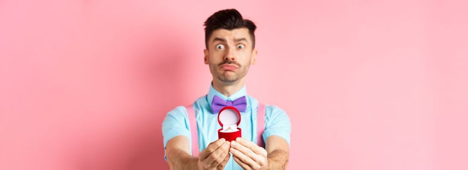 Valentines day. Nervous boyfriend waiting for girlfriend reply on marriage proposal, showing engagement ring and looking worried, standing over pink background.