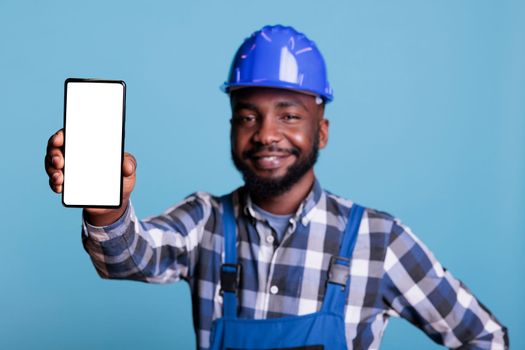 African american man holding cellphone with blank screen for advertising in front of camera. Professional builder wearing hard hat against light blue background, construction industry concept.