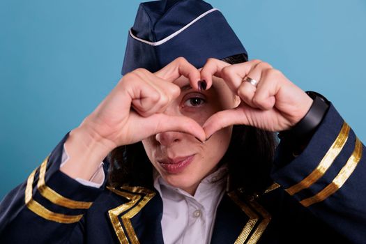 Smiling airliner stewardess showing heart shaped love symbol with fingers, conceptual romance gesture. Friendly air hostess in aviation uniform expressing affection feelings front view medium shot