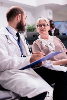 General practitioner filling out disability application for elderly patient at hospital lobby. Old woman answering routine questions from manager at sanatorium reception desk.