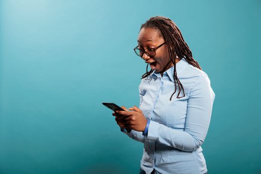 Cheerful excited african american person with smartphone device surprised by received email. Joyful smiling young woman with modern cellphone astonished by social media post reach.