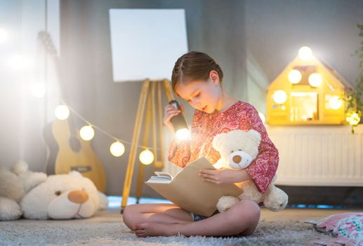 Little beautiful girl reading a book with flashlight in the bedroom. Child reads fairy tale with teddy bear and Christmas lights. Bedtime.