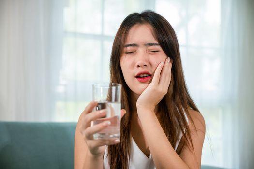 Asian young woman with sensitive teeth holding glass of cold water at home in living room, beautiful female toothache and dental problems touching cheek feeling pain after drinking cold water