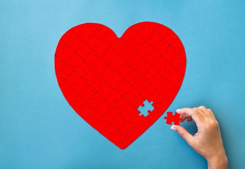 Red puzzle in heart shape. Hand with white details of puzzle on blue background.