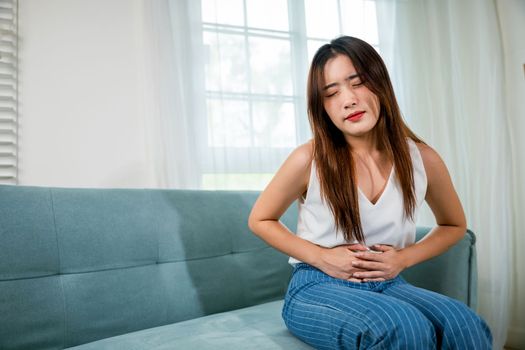 Sad Asian woman close eyes she problem painful stomachache, young female unhappy hands holding on stomach suffering from abdominal pain on sofa at home in living room, medical care and health concept