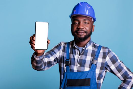 Construction worker in blue uniform work coveralls holds cell phone with empty screen for advertising isolated on blue background. Male professional builder in hard hat looking at camera.