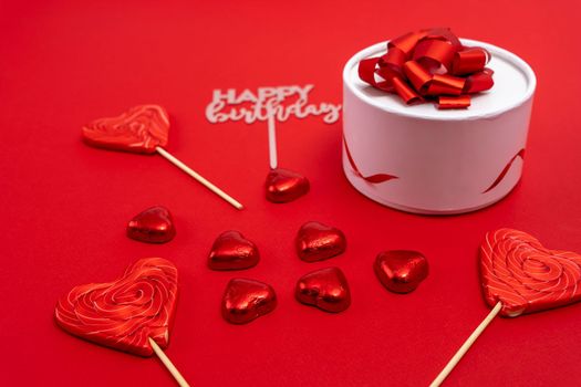 creative minimalist composition. a gift box with a ribbon and a bow, heart-shaped chocolates and heart-shaped caramel lollipops on a red background. A declaration of love. a gift and gratitude. Valentine's Day. postcard. copy space