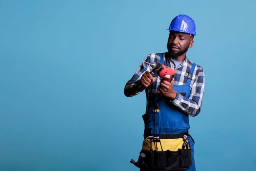 Builder with drill and belt with all tools doing renovation for client, drilling holes in the wall or doing renovations. Professional builder in hard hat with tool belt on light blue background.