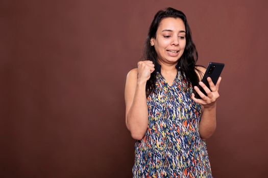 Excited indian woman showing winning gesture, reading good news, victory celebration. Amazed happy lady with clenched fist holding smartphone, looking at phone screen, celebrating success