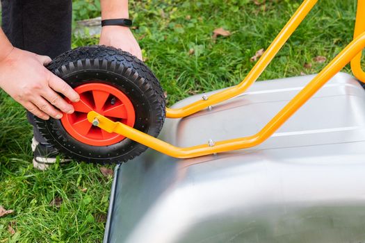 There is a metal wheelbarrow on the street, a man spins a new wheelbarrow for work, tightens bolts and nuts, hangs a wheel