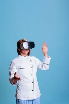 Confident elderly cook wearing virtual reality glasses while preparing culinary recipe working at healthy meal. Friendly chef with restaurant uniform cooking gastronomy food dish. Food industry