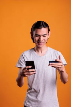 Smiling young man making order in internet shop, holding smartphone and credit card. Asian cheerful teenager buying products, searching goods in online store, e commerce concept