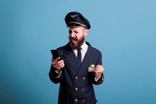 Happy smiling airplane pilot in uniform holding modern phone with winner gesture, clenched fist. Pilot with cheerful facial expression looking at mobile phone screen, reading good news