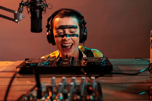 Woman performer acting silly at mixer, looking closely at control buttons to mix and remix music wearing headphones. Producing techno sounds with turntables, funky focused disc jockey.