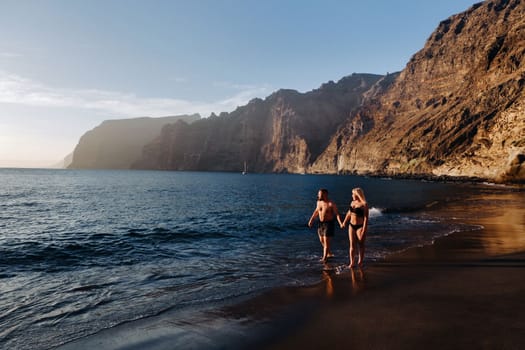 A couple in love walks on the beach against the backdrop of the Acantilados de Los Gigantes mountains at sunset, Tenerife, Canary Islands, Spain.