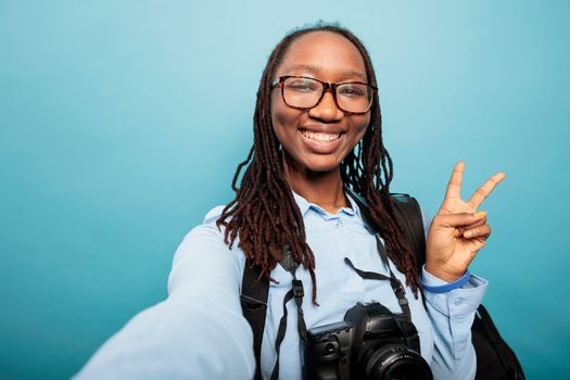 Positive happy photography entusiast with DSLR camera making peace sign with fingers while taking selfie photo. Confident optimistic young woman standing on blue background while gesturing victory.