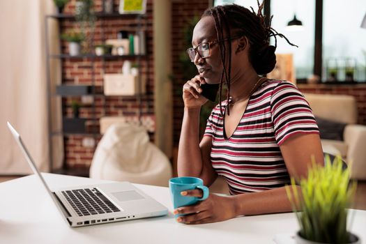 Freelancer talking on smartphone, working on laptop, drinking tea in home office. Busy remote worker answering phone call, employee having conversation on telephone, sitting at desk