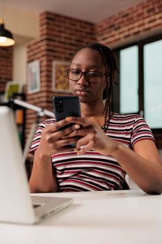 Freelancer sending text messages on smartphone, chatting in social network at workplace. Remote worker in glasses having break, surfing internet on mobile phone, sitting at desk