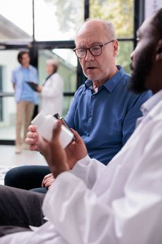 African american physician explaining medication treatment to senior patient while showing painkiller pills during checkup visit appointment in hospital waiting area. Medicine service and concept