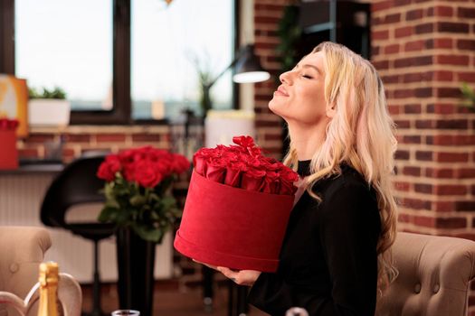 Woman holding valentine day roses in heart shaped box, standing in living room filled with romantic gifts. Seductive girlfriend enjoying celebrating love holiday, presents and flowers bouquet