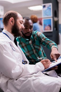 Caucasian general practitioner showing prescription to young african american sick man at hospital reception desk. Male patient giving information about medical condition to doctor in sanatorium.