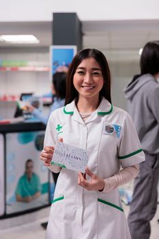 Smiling asian pharmacist holding box of vitamins in medicinal drugstore, showing package of medicaments and supplements. Advertising pills and drugs from pharmacy shop shelves.