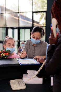 Mother standing at reception with child signing papers with medical insurance during appointment in hospital waiting area. People wearing protective face mask to prevent infection with coronavirus