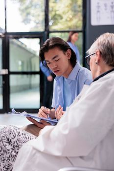 Young asian patient signing medical document while elderly doctor explaining health care treatment for curing disease. Multi ethnic people standing in hospital waiting area during consultation