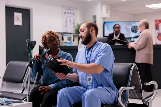 African american woman looking images of injuries on tablet. Physician at rehabilitation clinic explaining therapies to young patient with leg conditions. Sanatorium waiting room with diverse people.