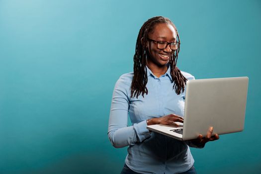 Cheerful happy woman having laptop and smiling heartily while browsing webpages on internet. Joyful pretty lady with handheld modern computer standing on blue background while surfing browser.