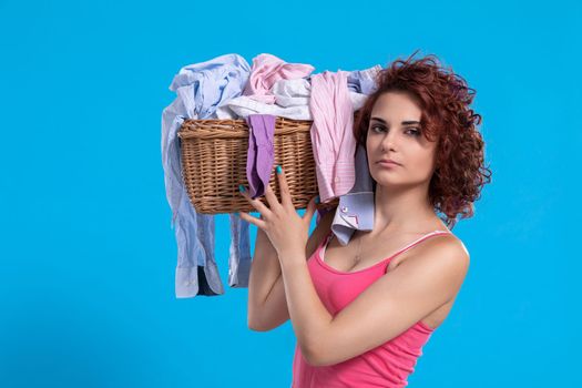 A strong woman holds a heavy wicker basket full of laundry clothes on her shoulder. An attractive girl with dark and curly hair.